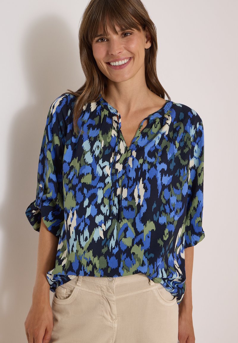 Femme souriante portant un chemisier ample à motifs bleus, verts et noirs avec les manches retroussées et un pantalon beige, les mains dans les poches.
