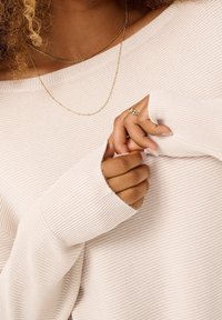 Maglione a maglia beige chiaro con maniche lunghe e flessibili. Presenta una texture a coste e collane di delicatezza dorate sovrapposte. Mani adornate con anelli.
