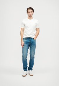 Tricou alb din bumbac, blugi din denim albastru cu ușoare estompari, și adidași albi cu talpă din cauciuc maroniu. Modelul stă cu mâinile în buzunare.