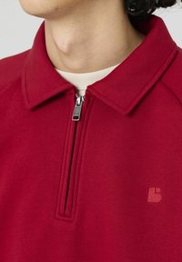 Sweatshirt zippé rouge avec un col, arborant un logo texturé sur la poitrine et une doublure intérieure contrastante. Tissu doux et lisse.