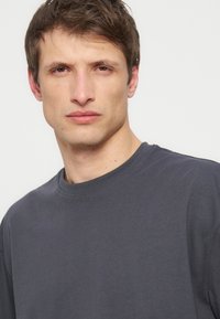 Νέος άνδρας με κοντά καστανά μαλλιά που φορά ένα απλό σκούρο γκρι μπλουζάκι με στρογγυλή λαιμόκοψη, κοιτάζοντας μπροστά, μπροστά σε ανοιχτό φόντο.