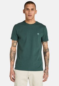 Tmavě zelené bavlněné tričko s kulatým výstřihem, krátkými rukávy a jemným logem na hrudi. Hladká textura, bez vzorů nebo akcentů.