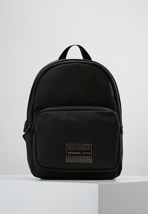 Sac à dos noir Versace Jeans avec poche zippée à l'avant, ornée d'une plaque logo cloutée en or, sur un fond clair uni.