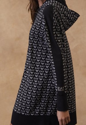 Zwarte hoodie-jurk met witte EA7- en adelaarlogo's herhaald, gedragen door een persoon die vanaf de zijkant wordt getoond, beige achtergrond.