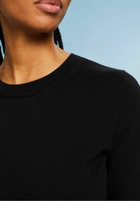 Gros plan d'une personne portant un pull ras du cou noir sur un fond bleu clair, montrant le cou, le bas du visage et l'épaule.