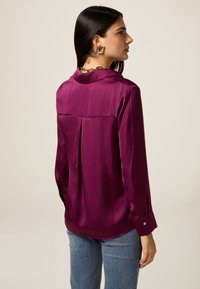 Paarse satijnen blouse met lange mouwen, een achterste plooien, en met knoopsluiting aan de manchetten, gecombineerd met blauwe denim jeans. Gladde textuur met een glanzende afwerking.