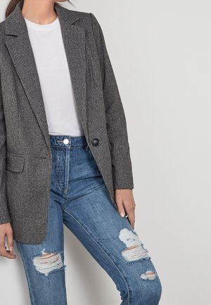 Femme portant un blazer gris à carreaux, un t-shirt blanc et un jean bleu déchiré, debout devant un fond clair uni.