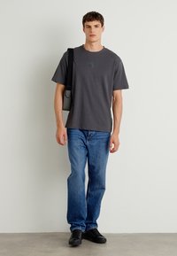 T-shirt en coton gris à manches courtes, assortie à un jean en denim bleu. Chaussures noires et sac à bandoulière noir. Se tenant contre un fond neutre.
