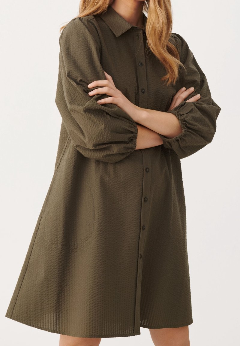 Femme portant une robe boutonnée texturée vert olive à longues manches bouffantes, debout les bras croisés.