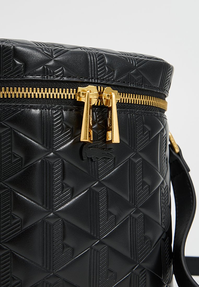 Sac noir rond avec motif géométrique embossé, fermeture éclair dorée et accents matériels dorés. Logo Lacoste présent sur le devant.