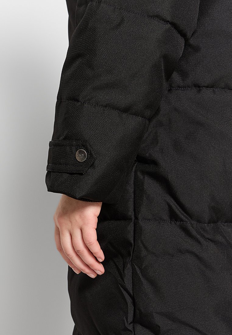 Veste en duvet noire avec un tissu texturé, un design rembourré et un détail de poignet à boutons. Gros plan d'une main reposant sur la manche.