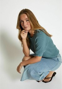 Tealfarbener Pullover aus Strick mit strukturiertem Muster, kurze Ärmel; kombiniert mit einem hellen Jeansrock und schwarzen Flip-Flops. Das Model hockt und stützt das Kinn auf die Hand.