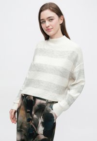 Tânără purtând un pulover în dungi albe și gri cu pantaloni largi, cu model, pozând cu o mână în buzunar, pe fundal alb.