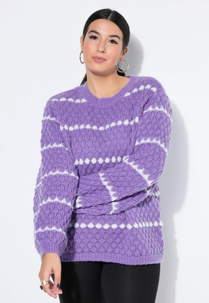 Tummahiuksinen nainen purppuranvioletti neulepaita, jossa valkoiset raidat, käyttää renkaanmuotoisia korvakoruja ja mustia housuja, seisoo valkoista taustaa vasten.