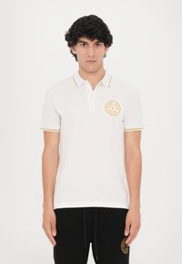 Bela polo majica z zlatimi detajli na ovratniku in rokavih. Ima okroglo logo na levem prsnem delu. Tkanina se zdi gladka in lahka.