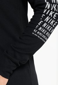 Schwarzes Langarmshirt aus strukturiertem Stoff mit gerippten Bündchen, das entlang des Ärmels mehrmals mit dem weißen "Nike"-Logo bedruckt ist.