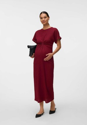 Femme enceinte portant une longue robe bordeaux, tenant une pochette noire et touchant doucement son ventre, avec des chaussures noires à talons bas.