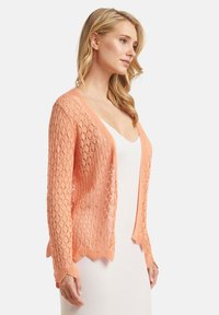 Luftige Strickjacke in hellem Pfirsich mit einem Rautenmuster, langen Ärmeln und welligem Saum. Leichte und halbtransparente Textur.