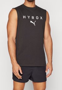 Musta hihaton urheilullinen tank-top valkoisella "HYROX"-tekstillä ja Puma-logolla. Sujuva puuvillamateriaali, istuva muotoilu, minimalistinen tyyli.