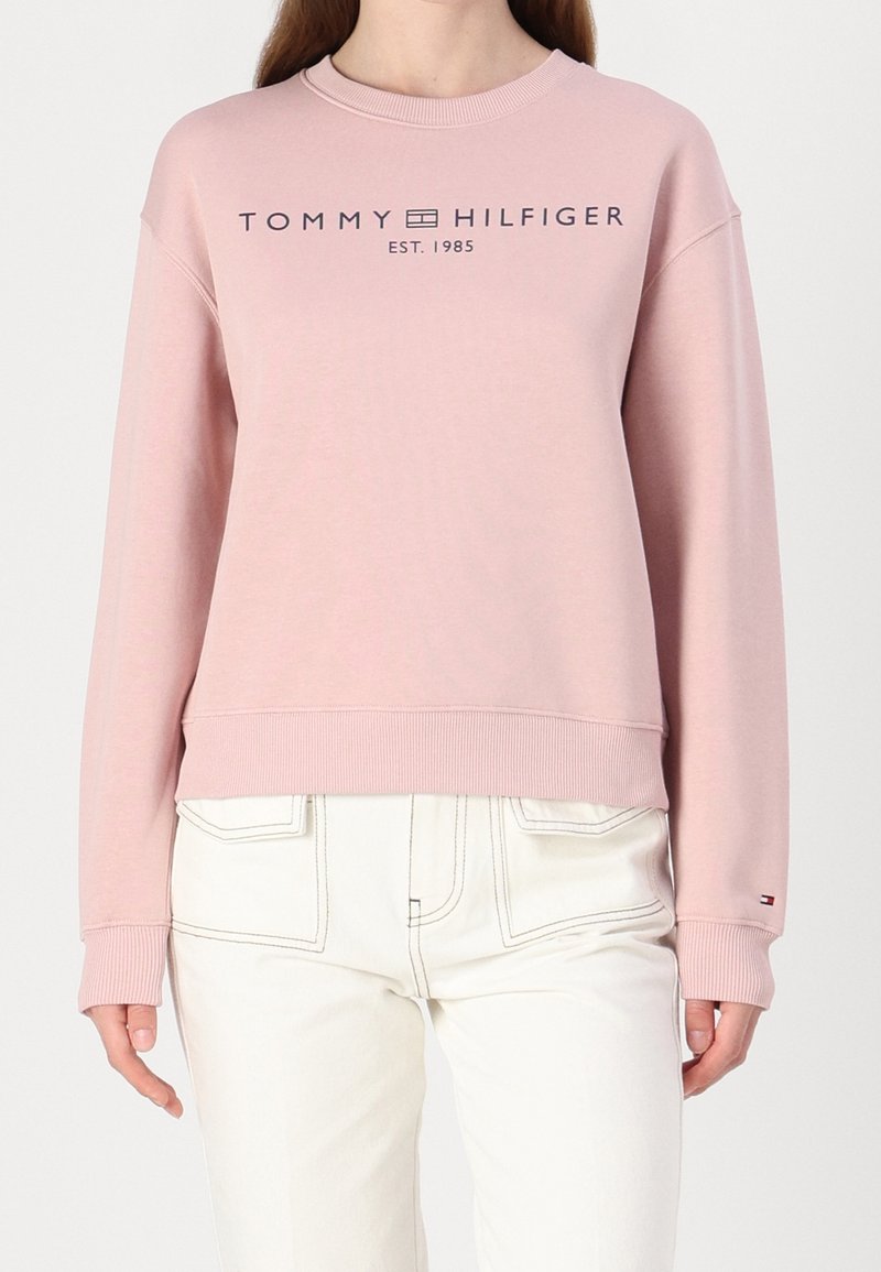 Personne portant un sweat-shirt rose clair Tommy Hilfiger avec logo et texte "EST. 1985", assorti à un pantalon blanc taille haute.
