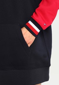 Marinefarbenes Sweatshirt mit roten Ärmeln, ausgestattet mit einer Fronttasche, gerippten Bündchen in Schwarz, Weiß und Rot sowie einem Logodetail am Ärmel.