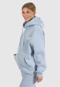 Felpa oversize di colore azzurro chiaro realizzata in morbido cotone misto, con cordoncini, tasche frontali e un piccolo logo stampato sul petto.