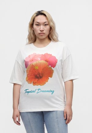 Jeune personne aux longs cheveux blonds porte un t-shirt blanc avec de grandes fleurs d'hibiscus orange et rose et le texte « Tropical Dreaming », jean bleu clair.