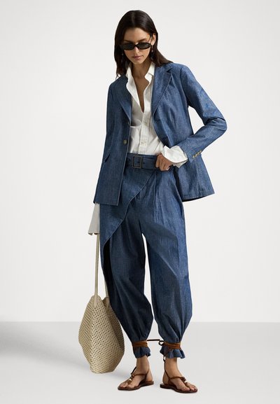 Džínový oblek s výrazným blejzrom a voľnými nohavicami, doplnený o bielu košeľu, svetlohnedé sandále a pletenú béžovú tašku. Tmavomodrá farba.
