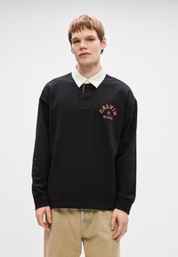 Μαύρο μακρυμάνικο πόλο μπλουζάκι με λευκό γιακά, με κόκκινο κείμενο "CALVIN KLEIN" μπροστά, φτιαγμένο από απαλό ύφασμα. Χαλαρή εφαρμογή.