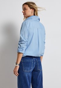 Ljusblå, långärmad skjorta med knappar och uppvikta ärmar, detalj med veck bak, kombinerad med mörkblå denimjeans med bakfickor.