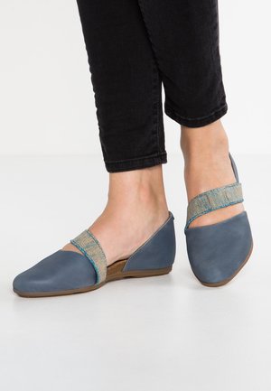 Femme portant des chaussures plates bleu slip-on avec une sangle élastique beige texturée et un pantalon noir raccourci sur un sol blanc.