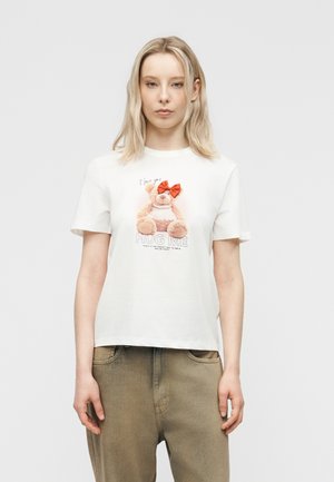 Νεαρή γυναίκα που φοράει λευκό μπλουζάκι με γραφικό αρκουδάκι, κόκκινο φιόγκο και κείμενο «Αγκαλιάστε με», συνδυασμένο με μπεζ παντελόνι, στέκεται μπροστά σε απλό φόντο.