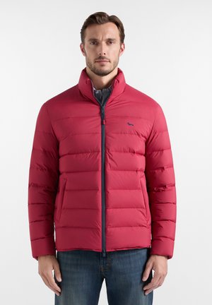 Uomo che indossa una giacca trapuntata rossa con collo alto e zip frontale, abbinata a jeans blu, in piedi davanti a uno sfondo semplice.
