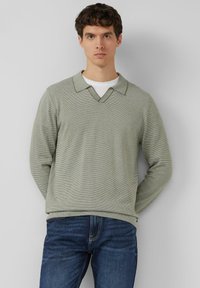 Lichtgroene trui met lange mouwen, V-hals en een subtiel streeppatroon, gecombineerd met donkerblauwe spijkerbroek.