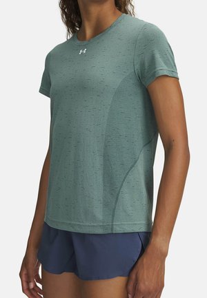 Frau trägt grünes kurzärmeliges Sportshirt mit einem Logo auf der Brust und marineblaue Laufshorts, steht vor einem einfarbigen Hintergrund.