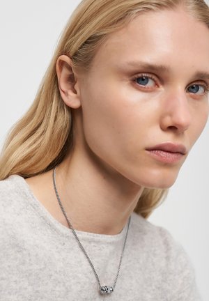 Silberne Kettenhalskette mit einem runden Anhänger. Das Model hat hellblonde Haare und trägt einen grauen, strukturierten Pullover. Nahaufnahme des Gesichts.