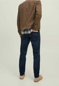 Giacca in suede marrone, camicia a quadri, jeans blu scuro e scarpe beige chiaro. La giacca ha un design cropped e bottoni in vita.