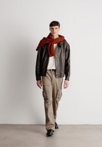 Pruun nahktagi valge särgi peal, oranž sall kaela ümber, khaki kargo püksid ja mustad kingad. Neutraalne taust.