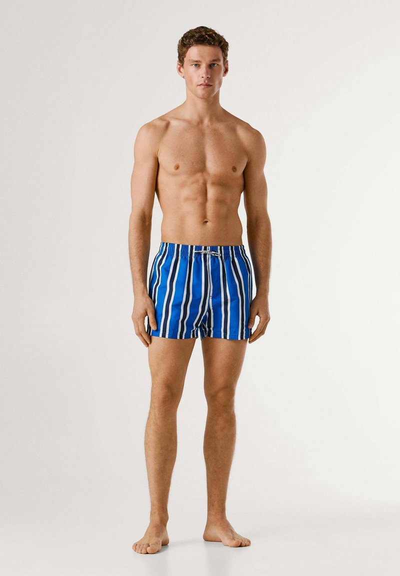 Jeune homme debout pieds nus portant un short de bain rayé vertical bleu et blanc sur un fond uni.
