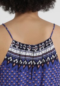 Detailní záběr zadní části horní části těla osoby, která má na sobě modrou šatovku bez rukávů s tenkými ramínky a klíčovou skulinkou uprostřed.