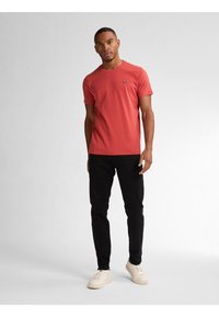 Rode T-shirt met korte mouwen en een ronde halslijn, voorzien van een klein logo op de borst. Gekleed met zwarte broek en lichte sneakers.