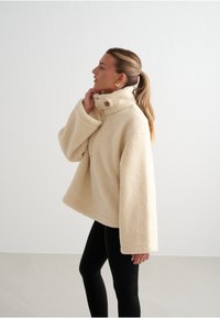 Krémová fleecová bunda s vysokým límcem, oversize střihem a velkými manžetami. Obsahuje zapínání na knoflíky a má jemnou strukturu.