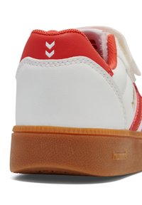 Weißer Sneaker mit roten Akzenten, gebogenem Absatz, strukturiertem Gummisohle und kontrastierenden Nähten. Verfügt über ein Logo und ein Chevron-Detail auf der Rückseite.