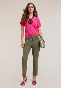 Różowa bluzka z krótkim rękawem i bufkami, oliwkowe spodnie, różowe sandały na obcasie oraz torebka w wzory. Zawiera także czarną naszyjnik-paretę.