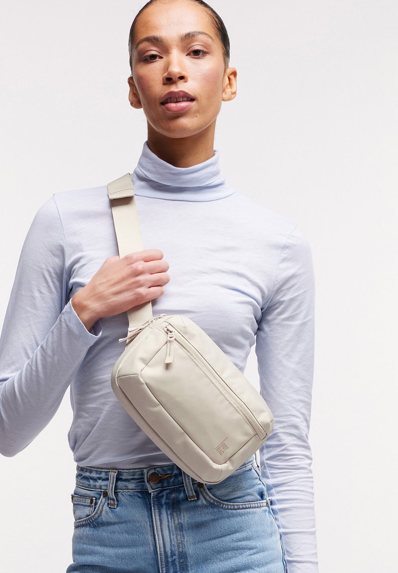 Femme portant un col roulé bleu clair et un jean, tenant un sac bandoulière beige avec une large sangle sur un fond uni.