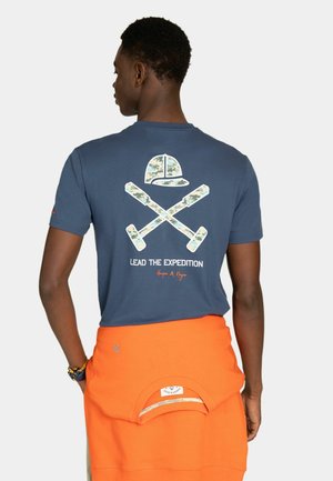 Hombre con camiseta azul con gráfico de palas de camuflaje cruzadas y gorra, y texto "Lead the Expedition", sudadera naranja atada a la cintura.