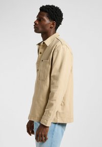 Camisa button-up color beige con un bolsillo frontal, mangas largas y un corte relajado. Fabricada con un material resistente, cuenta con un cuello y detalles cosidos.