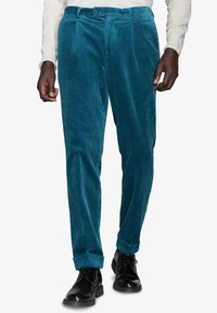 Teal corduroy-housut, joissa on ribbikuvio, edessä laskokset, nappikiinnitys ja käännetyt lahkeet, käytössä mustien kenkien kanssa.