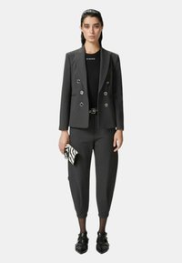 Blazer grigio su misura con bottoni doppi sopra un top nero, abbinato a pantaloni grigi a sigaretta e una pochette con fantasia. Scarpe nere completano il look.