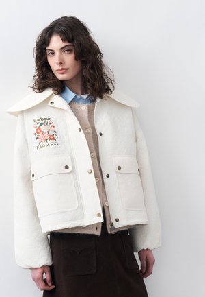 Γυναίκα με καστανά σγουρά μαλλιά που φοράει λευκό καπιτονέ μπουφάν με λουλουδάτο σχέδιο, μπεζ ζακέτα, μπλε πουκάμισο και σκούρα φούστα μπροστά από ένα απλό τοίχο.